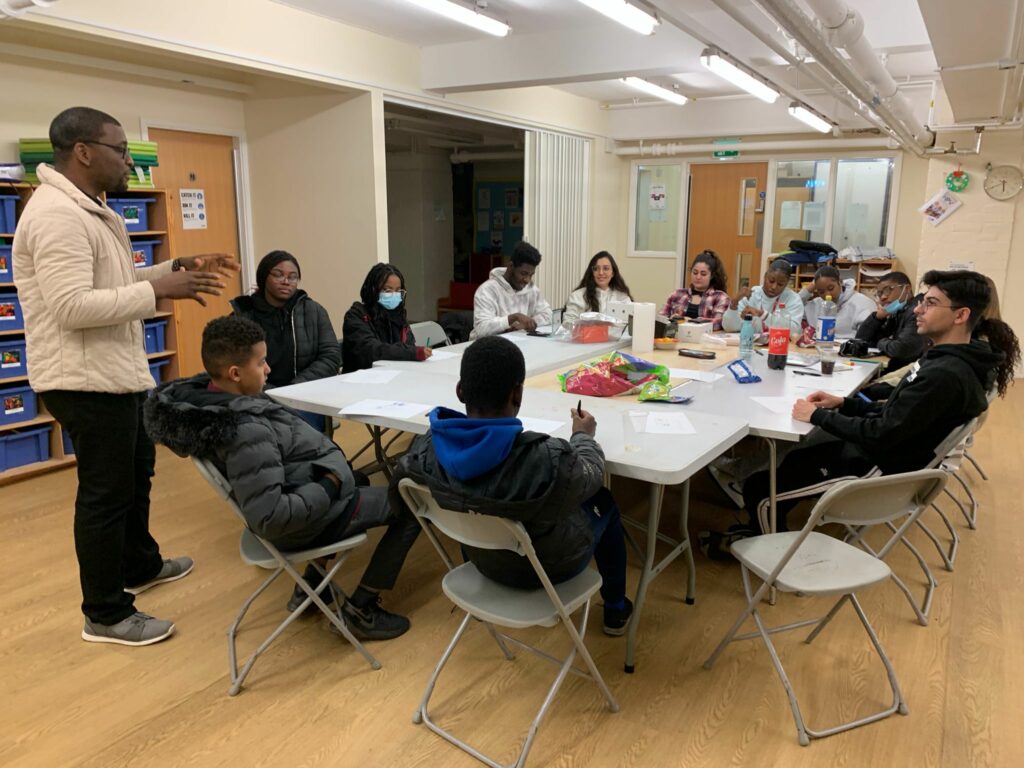 STAND4 youth group convene around a table with workshop host, Demola presenting at the front