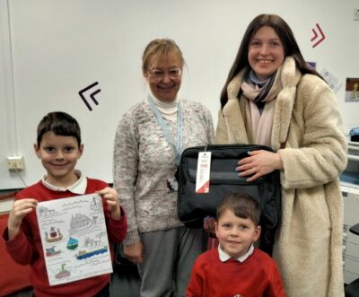 Digital divide donation. Iryna receives a laptop donation from Success4All. Two young brothers wearing a red jumper smile, with the older of the two holding a drawing on different boats. 